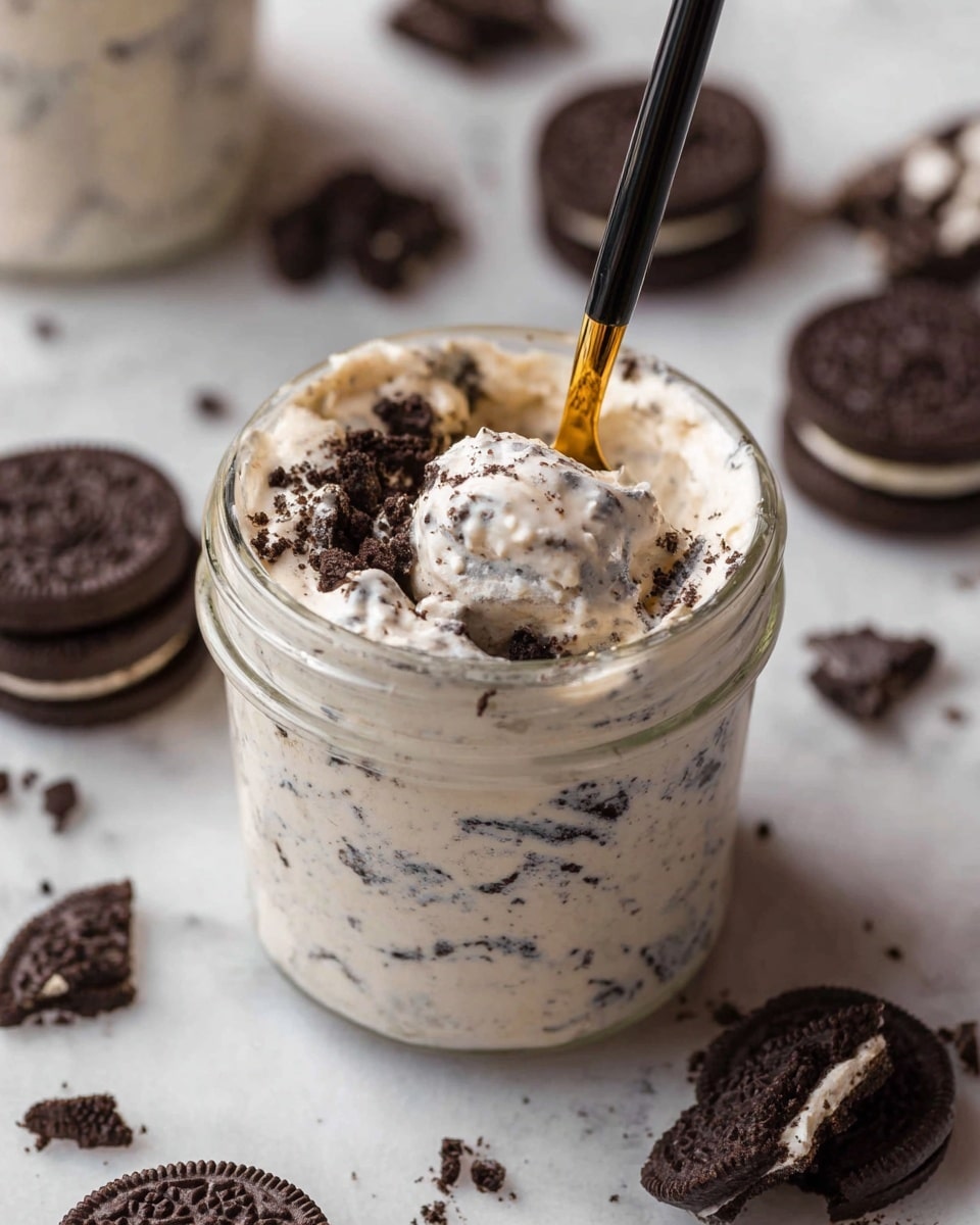 The image shows a clear glass jar filled with a creamy white dessert mixed with small dark cookie pieces, giving it a speckled look. Inside the jar, a spoon with a black handle and gold end is dipped into the dessert, showing its soft and fluffy texture. Around the jar, there are whole and broken dark chocolate sandwich cookies placed on a white marbled surface. The dessert fills the jar nearly to the top, and the cookie pieces inside stand out against the light cream, making it look rich and textured. photo taken with an iphone --ar 4:5 --v 7