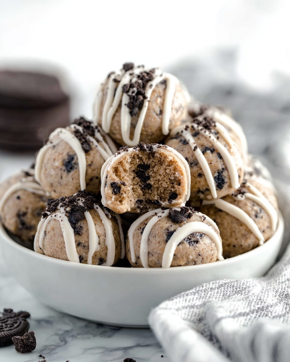 A white bowl filled with round cookie dough balls that are light brown with small dark cookie pieces inside and on top. Each ball is drizzled with thin white icing lines from the top, creating a striped pattern, and topped with dark cookie crumbs. One cookie dough ball is broken open, showing a dense, textured inside with small dark cookie pieces. The bowl is placed on a white marbled surface, with a soft white and grey striped cloth and some dark cookies blurred in the background. photo taken with an iphone --ar 4:5 --v 7