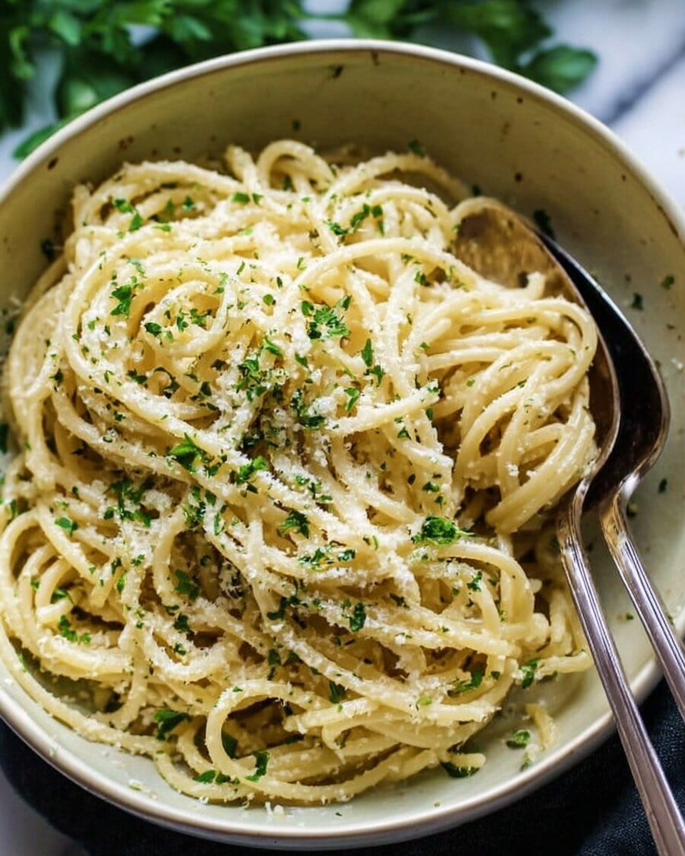 A white bowl filled with spaghetti noodles coated in a light creamy sauce, sprinkled with finely chopped green herbs and grated cheese on top. Two silver spoons rest inside the bowl on the right side. The bowl sits on a white marbled surface with some green leaves softly visible in the blurred background. The noodles look soft and twisted, creating a textured, inviting appearance. photo taken with an iphone --ar 4:5 --v 7