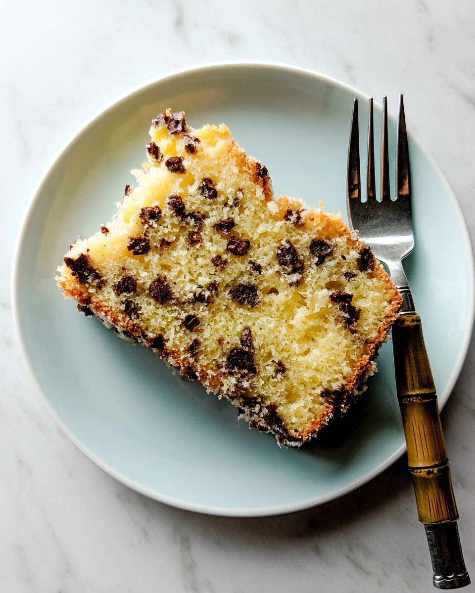 A single slice of chocolate chip cake with a light golden-yellow inside and small dark brown chocolate chips spread evenly throughout, placed on a white plate. The cake has a rough textured top edge with slightly browned bits. On the plate next to the cake, there is a silver fork with a bamboo-like handle resting diagonally. The whole setting is on a white marbled surface. photo taken with an iphone --ar 4:5 --v 7