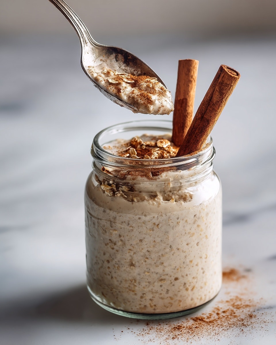 A clear glass jar filled with layered oatmeal that has a creamy beige color with small bits visible inside, topped with a sprinkling of cinnamon powder giving a light brown dusting on the surface, and two whole cinnamon sticks standing upright on one side. A shiny silver spoon is held above the jar, scooping a portion of the oatmeal that looks soft and thick with sprinkled cinnamon and oats clinging to it. The jar is set on a white marbled surface with blurred soft background colors. Photo taken with an iphone --ar 4:5 --v 7