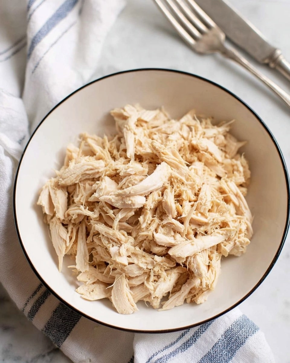A white bowl with a thin black line around the edge is filled with shredded light beige cooked chicken. The chicken pieces are uneven in size and texture, some thin and stringy, others slightly thicker. The bowl is set on a white marbled surface next to a white cloth with blue stripes, and an old silver fork and knife are placed nearby. The lighting is soft and natural, showing the detail and slight gloss of the chicken. photo taken with an iphone --ar 4:5 --v 7