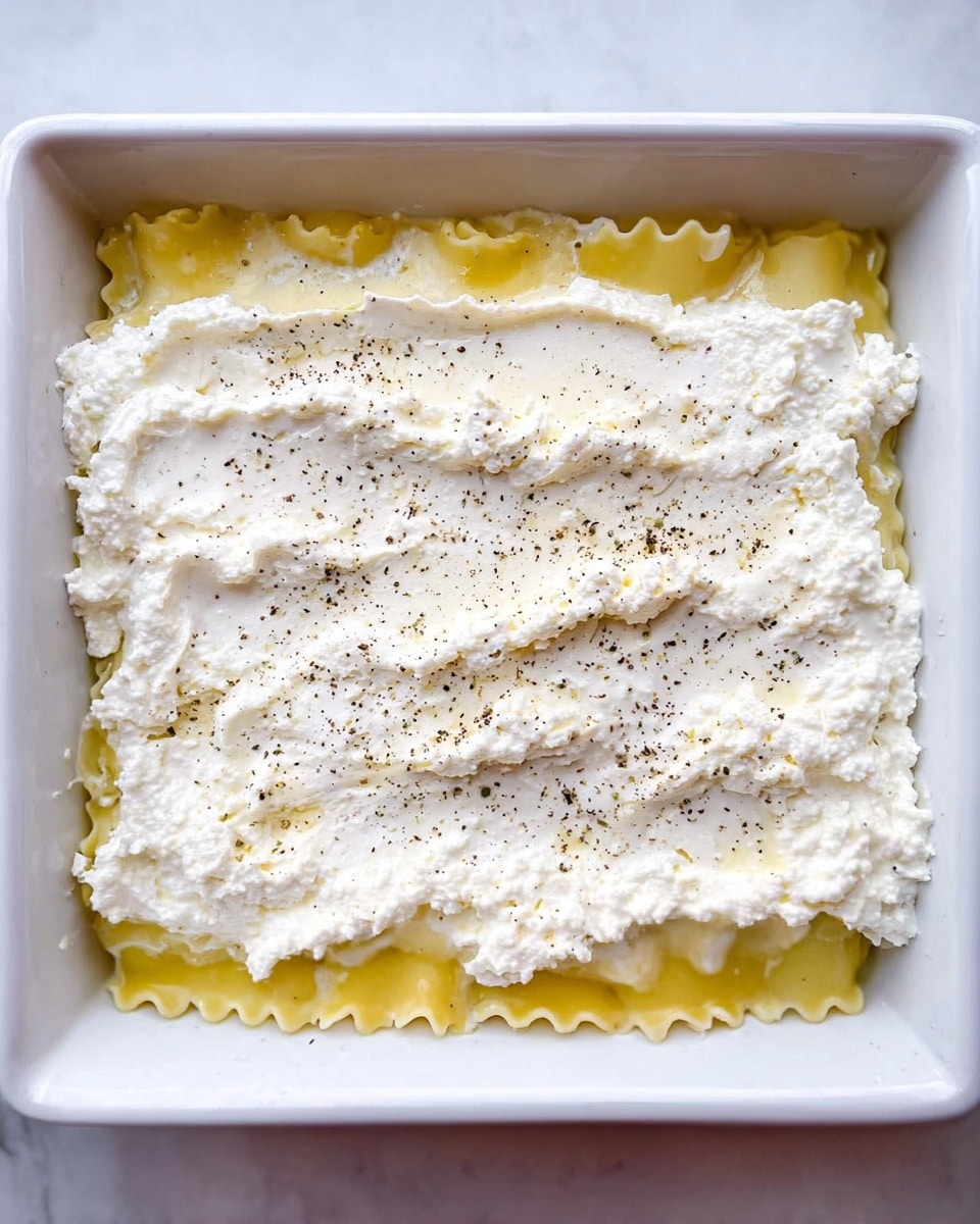 The image shows a white rectangular baking dish on a white marbled surface, filled with one layer of yellow lasagna pasta at the bottom. On top of the pasta, there is a thick, uneven layer of white ricotta cheese spread across, with some small holes and texture visible. A light sprinkling of black pepper is scattered on the cheese, adding small dark specks. The overall look is fresh and simple, with soft and slightly rough textures. Photo taken with an iphone --ar 4:5 --v 7