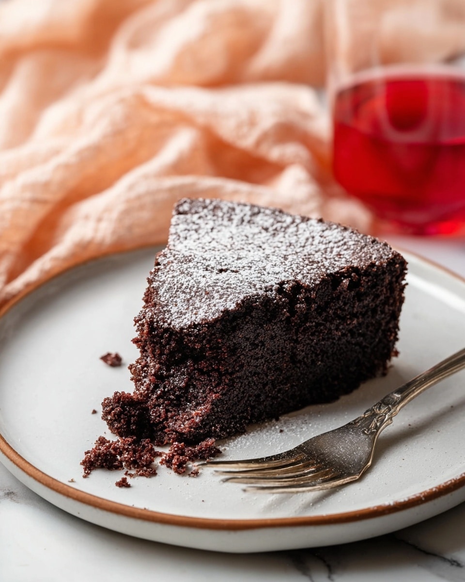 A single thick slice of dark chocolate cake with a rich, moist texture sits on a white plate with a thin brown rim. The top layer is dusted lightly with white powdered sugar, creating a soft contrast against the dark chocolate surface. The dense cake has an uneven, slightly crumbly edge, showing its moist inside. Next to the slice on the plate, a silver fork rests with its tines pointed towards the cake. In the very front, slightly out of focus, a glass with bright red liquid is visible. The background has a soft peach cloth draped gently over a white marbled surface. photo taken with an iphone --ar 4:5 --v 7