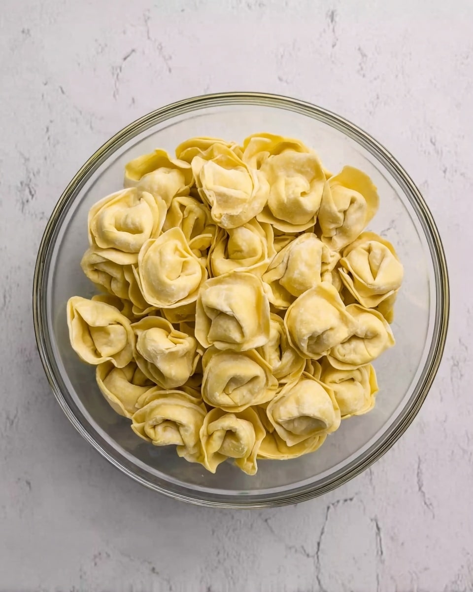 A clear glass bowl filled with many pale yellow tortellini pasta pieces, each showing a folded shape with a slightly rough texture. The bowl is placed on a white marbled surface with soft grey veins, creating a clean and light background. The tortellini are arranged loosely and fill the bowl almost to the top, with some pieces overlapping slightly but mostly flat. The pasta has a matte finish with natural shadows highlighting its curves and folds. photo taken with an iphone --ar 4:5 --v 7