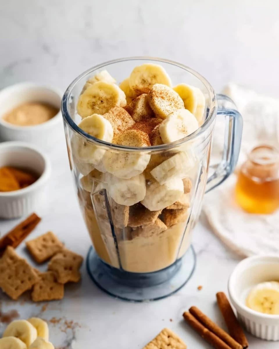 The image shows a clear blender filled with layers of sliced yellow bananas on top, light brown powder sprinkled over the bananas, and square pieces of light brown cereal or biscuit at the bottom. The blender is placed on a white marbled surface with a few sticks of cinnamon and sliced bananas nearby. In the background, there are small white bowls with a golden syrup and another light-colored ingredient, creating a cozy and fresh kitchen scene. Photo taken with an iphone --ar 4:5 --v 7