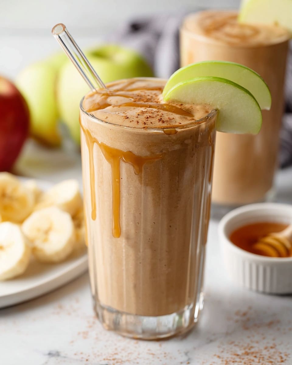 A tall clear glass filled with a light brown smoothie that has a creamy and smooth texture, topped with a drizzle of caramel-colored sauce and a sprinkle of fine cinnamon powder. Two thin slices of green apple rest on the rim of the glass, standing upright. A clear straw sticks out from the glass’s left side. The background shows blurred elements including a second glass with the same smoothie, apple slices, banana slices on a white plate, and a small white bowl filled with honey, all on a white marbled surface. Photo taken with an iphone --ar 4:5 --v 7