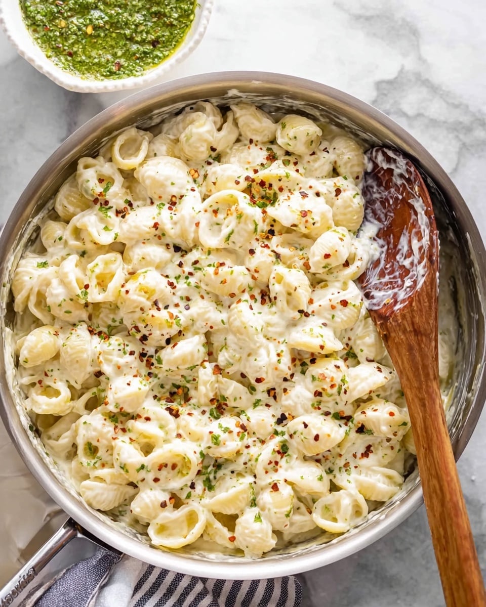 A silver pan filled with creamy white pasta shells covered in a thick white cheese sauce, with small green herb pieces and red pepper flakes sprinkled on top for color. A large wooden spoon rests in the pan, slightly coated with the sauce, and a white marbled surface is visible underneath. In the top corner, there is a white bowl with a green herb sauce inside. A soft striped cloth is partially visible under the pan. photo taken with an iphone --ar 4:5 --v 7