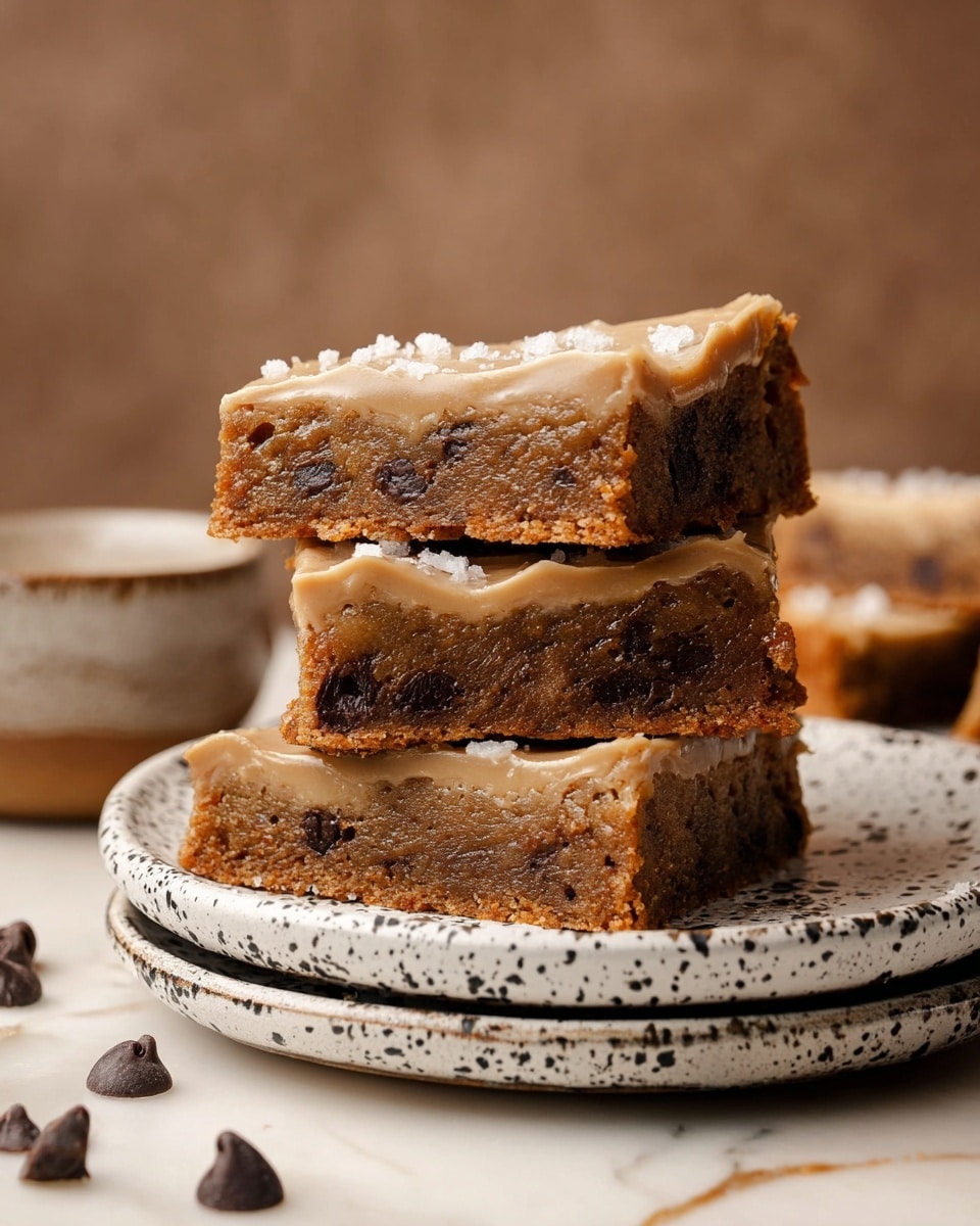 A stack of four chocolate chip cookie bars is shown on a white plate with black speckles, placed on two more similar plates. Each bar has a thick, dense, and chewy brown cookie layer filled with dark brown chocolate chips. On top of each bar is a smooth, light beige frosting with a slightly shiny and creamy texture, decorated with small flakes of white sea salt. Around the plate are a few loose chocolate chips scattered on a white marbled surface. The background is a soft brown blurred texture. photo taken with an iphone --ar 4:5 --v 7