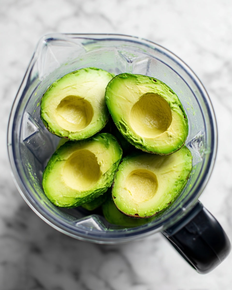 The image shows a clear blender container filled with four halves of green avocados with a soft, creamy texture inside. The avocados are bright green with some darker green near the skin. The blender is placed on a white marbled surface, and the black handle of the blender is visible on the top right side. photo taken with an iphone --ar 4:5 --v 7